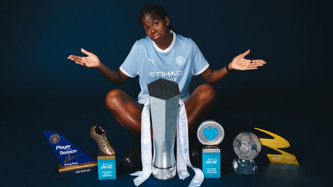 A footballer in a Manchester City kit sits on the floor with several trophies including Player of the Season and Golden Boot around them.