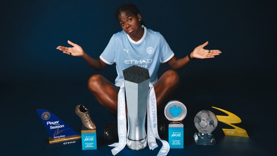 A footballer in a Manchester City kit sits on the floor with several trophies including Player of the Season and Golden Boot around them.