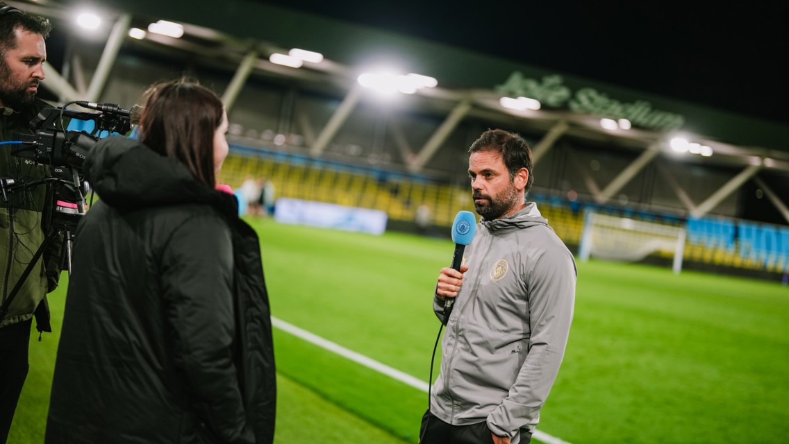 A man holding a microphone, speaking to a woman while another man operates a camera on a soccer field at night.