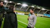 A man holding a microphone, speaking to a woman while another man operates a camera on a soccer field at night.