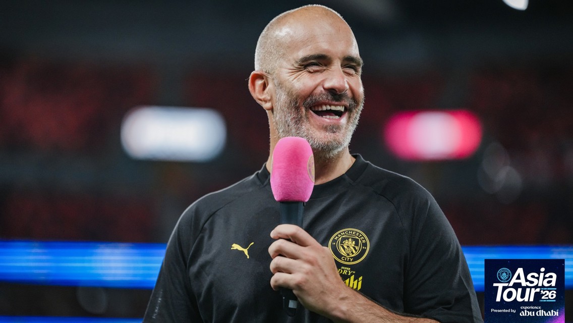 A man in a Manchester City shirt, holding a pink microphone and laughing during the Asia Tour '26 event.