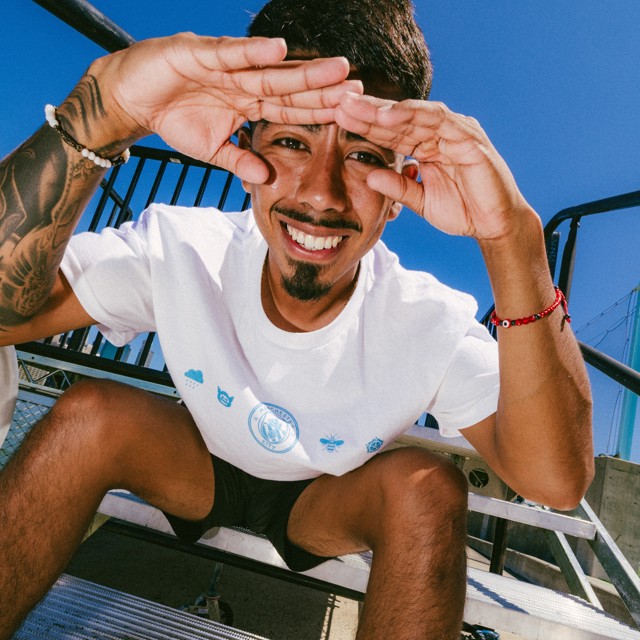 A person smiling while sitting on outdoor stairs under a bright blue sky, shading their eyes with their hands.