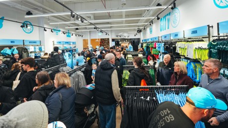 A crowded Manchester City merchandise store with people shopping for clothing and accessories. The store features sections for men, women, and junior items, including jerseys and other gear.