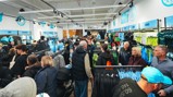 A crowded Manchester City merchandise store with people shopping for clothing and accessories. The store features sections for men, women, and junior items, including jerseys and other gear.