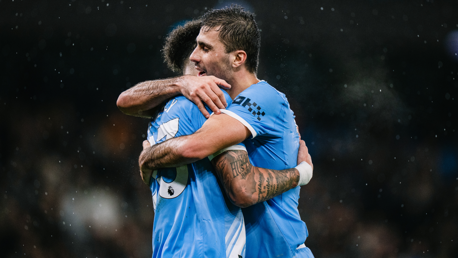 Two players in blue Manchester City jerseys hugging during a match celebration on a rainy sports field.