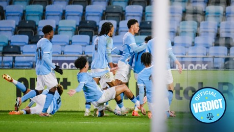 Manchester City players celebrate a goal on the field. Most are standing and embracing, while one player is sliding on his knees. Another player appears to be on the ground with his legs in the air. The scene is captured inside a stadium.