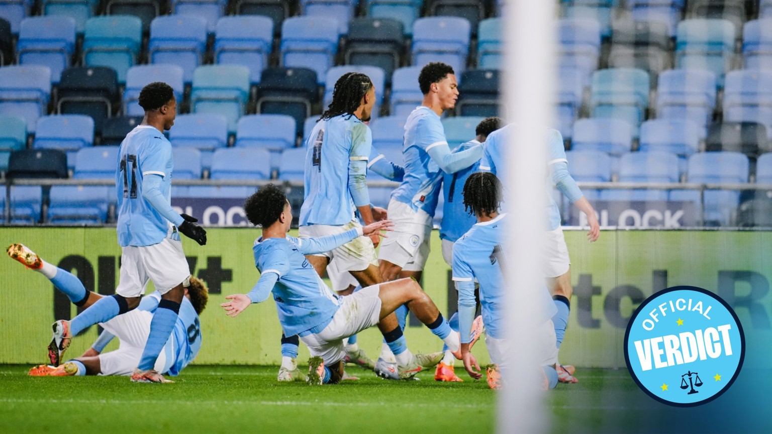 Manchester City players celebrate a goal on the field. Most are standing and embracing, while one player is sliding on his knees. Another player appears to be on the ground with his legs in the air. The scene is captured inside a stadium.