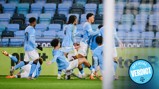 Manchester City players celebrate a goal on the field. Most are standing and embracing, while one player is sliding on his knees. Another player appears to be on the ground with his legs in the air. The scene is captured inside a stadium.