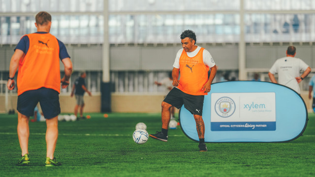 Two individuals wearing orange bibs during a football training session on an indoor pitch with a Manchester City sign and footballs visible.