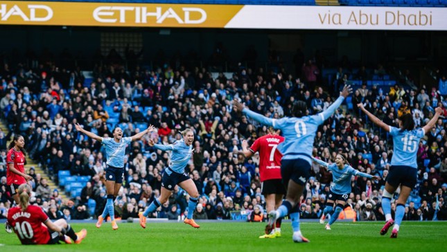 Manchester City Women's team celebrates a goal at Etihad Stadium with enthusiastic crowd. Players in light blue jersey, opposing team in red. Visible Etihad advertisement.