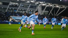 Manchester City players in action, wearing the blue kit, running on the soccer field during a match under stadium lights.