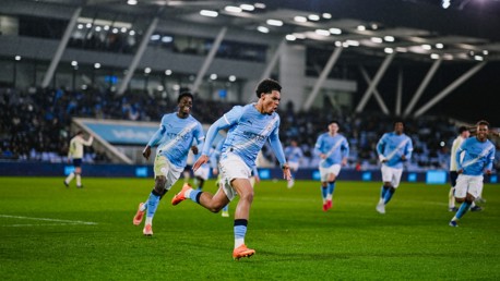Manchester City players in action, wearing the blue kit, running on the soccer field during a match under stadium lights.