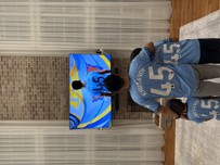A family of three, wearing matching Manchester City jerseys with the name Khusanov and number 45, watch a football match on television. The scene is set in a living room with wooden floors and brick wall.