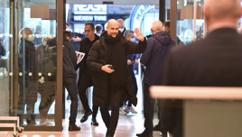 People walking through an entrance with Manchester City logo visible in the background. A man in the foreground is waving while holding a phone. Faces are blurred.