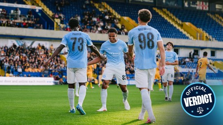 Manchester City youth team players celebrating a goal in blue jerseys with numbers 73, 83, and 98. Crowd and stadium visible in the background.
