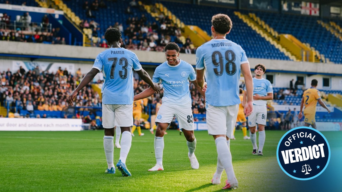 Manchester City youth team players celebrating a goal in blue jerseys with numbers 73, 83, and 98. Crowd and stadium visible in the background.