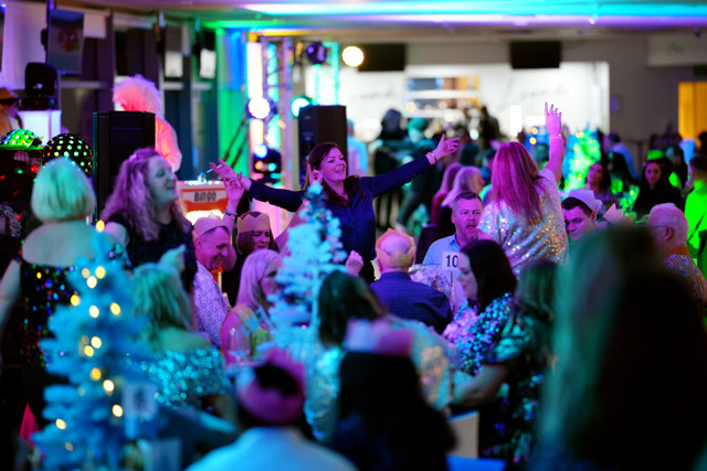 A vibrant scene at a party or event with people wearing colorful and glittery outfits, surrounded by festive lights and decorations, including mini Christmas trees.