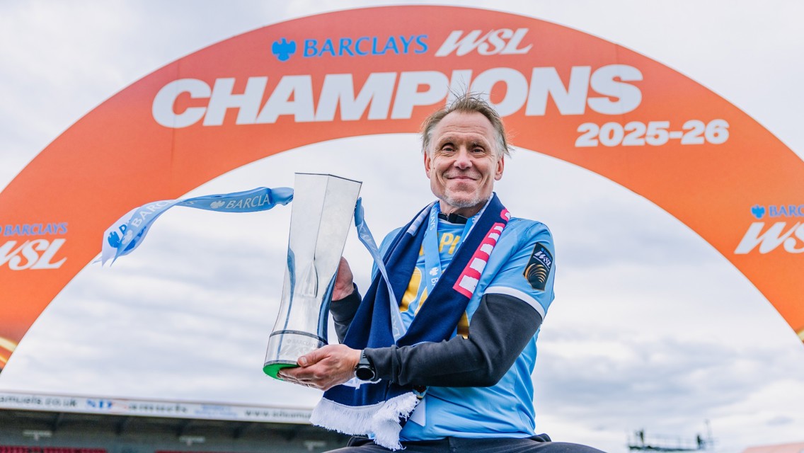 Person holding a trophy under an arch labeled 'BARCLAYS WSL CHAMPIONS 2025-26'.