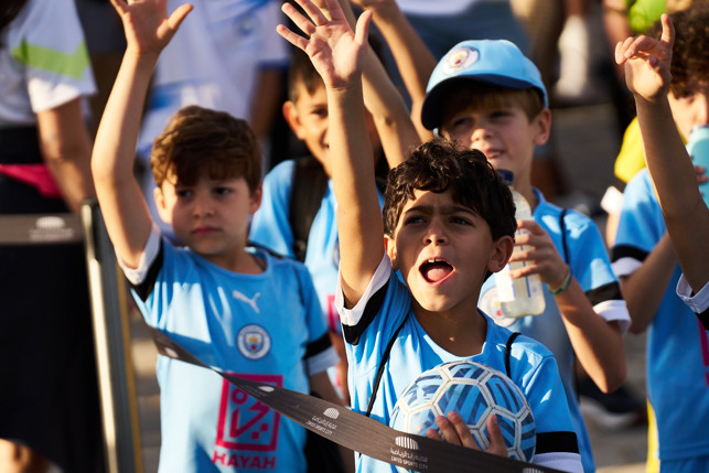 Several children wearing Manchester City jerseys are seen raising their hands. One child is holding a soccer ball, and another child is wearing a Manchester City cap. The image is taken at Zayed Sports City.