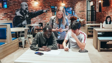Three individuals collaborating on a project in a cafe with exposed brick walls. Two people are seated at a table with papers and smartphones, while another stands over them with a lanyard and badge. Other patrons are seated in the background.