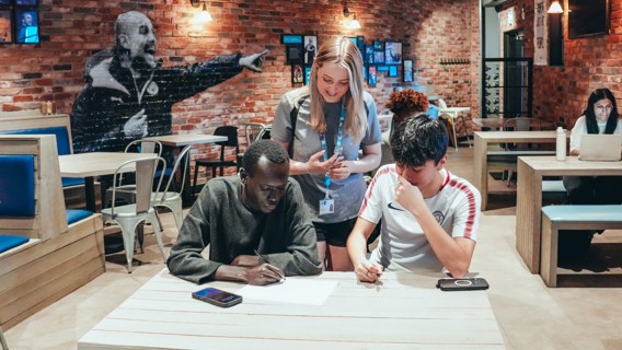 Three individuals collaborating on a project in a cafe with exposed brick walls. Two people are seated at a table with papers and smartphones, while another stands over them with a lanyard and badge. Other patrons are seated in the background.