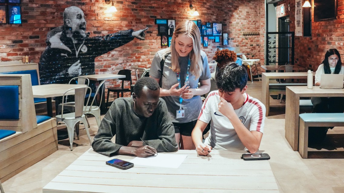 Three individuals collaborating on a project in a cafe with exposed brick walls. Two people are seated at a table with papers and smartphones, while another stands over them with a lanyard and badge. Other patrons are seated in the background.
