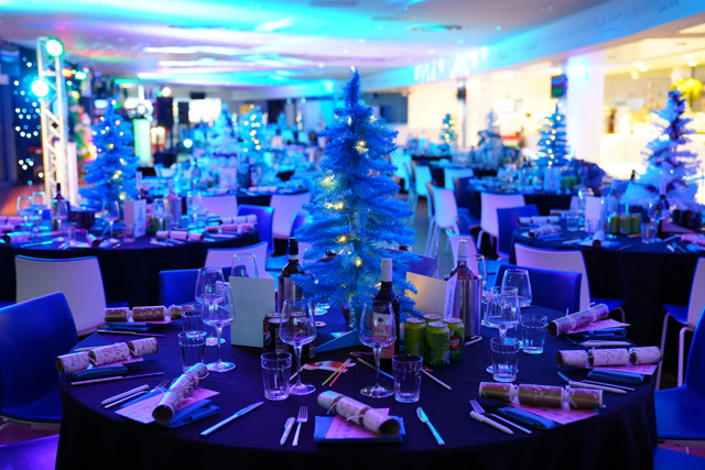 A festive gathering with tables set for dining. Each table features a small, lit Christmas tree centerpiece surrounded by drink bottles and glasses, cutlery, and crackers, under blue lighting.