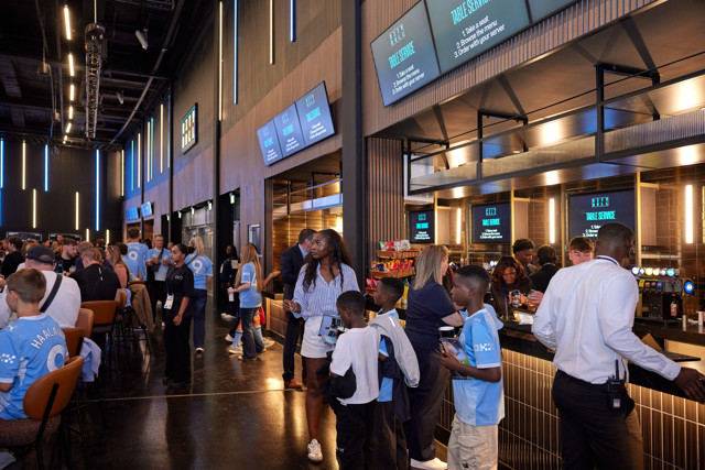 Guests enjoy food and drink at a concession stand inside a stadium lounge, surrounded by digital menu boards and wearing Manchester City jerseys.