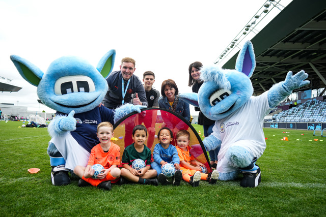 A group photo of children holding footballs with two Manchester City mascots and several adults at a community event on a football field.