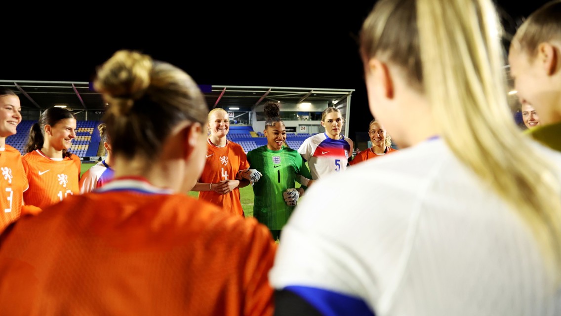 Football players gathered in a circle on the field. Some are wearing orange kits, and one is in a green goalkeeper outfit.