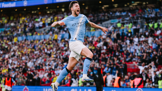 A Manchester City player celebrating a goal at a stadium filled with fans. The player is wearing the team's blue kit and jumping in the air with arms outstretched.