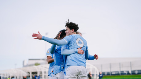 Football players in light blue jerseys celebrating a goal on the field. The jersey number 9 is visible on one player's back as they embrace and celebrate.