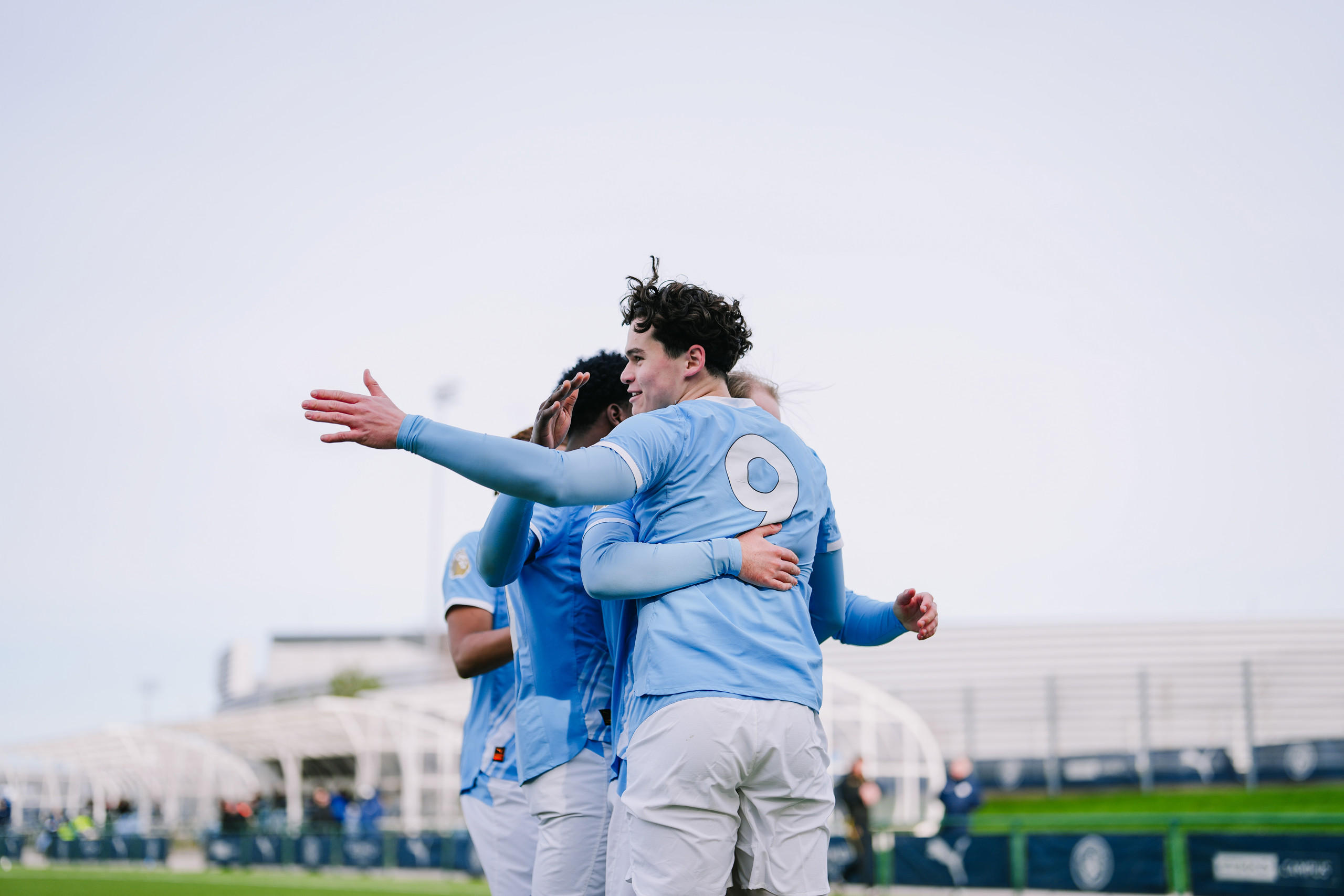Football players in light blue jerseys celebrating a goal on the field. The jersey number 9 is visible on one player's back as they embrace and celebrate.