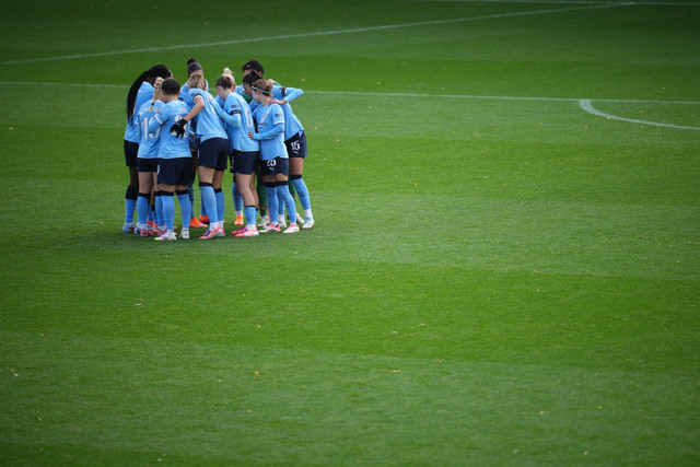 A group of football players in light blue uniforms huddling on a green soccer pitch, showcasing teamwork and unity.