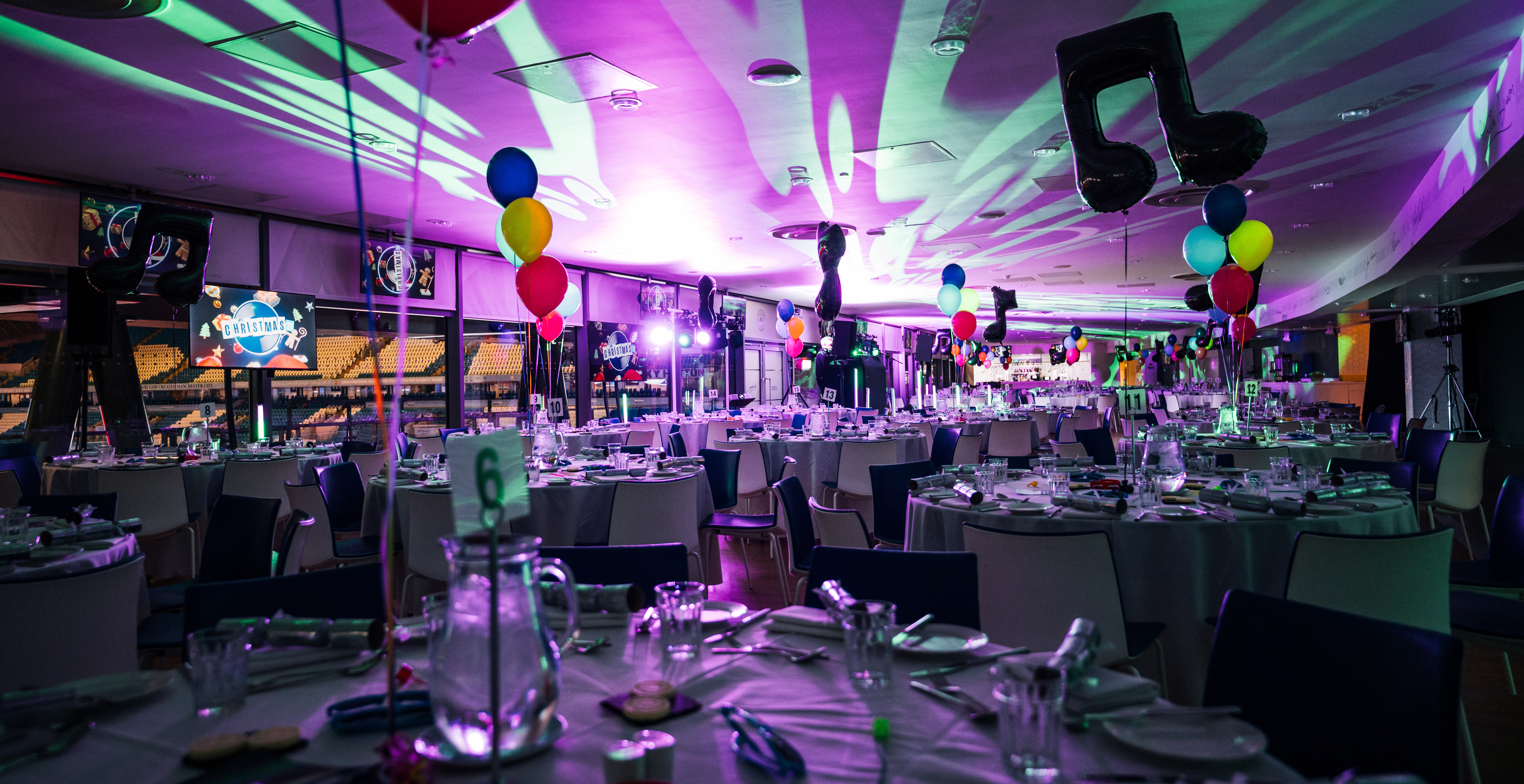 A festive venue with tables set for dining, decorated with colorful balloons, music-themed decorations, and Christmas imagery projected on screens.