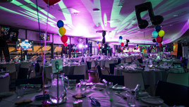 A festive venue with tables set for dining, decorated with colorful balloons, music-themed decorations, and Christmas imagery projected on screens.