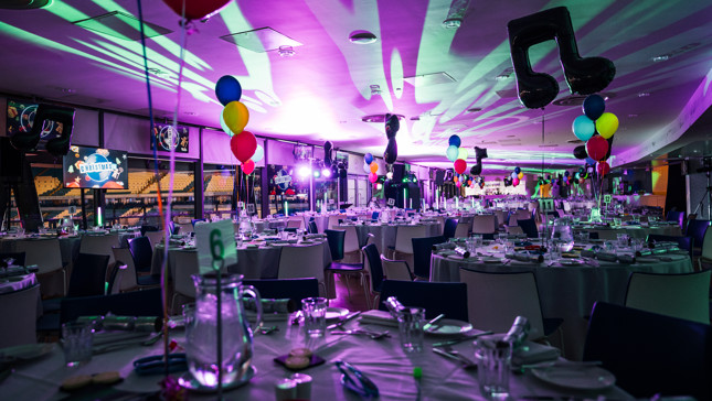 A festive venue with tables set for dining, decorated with colorful balloons, music-themed decorations, and Christmas imagery projected on screens.