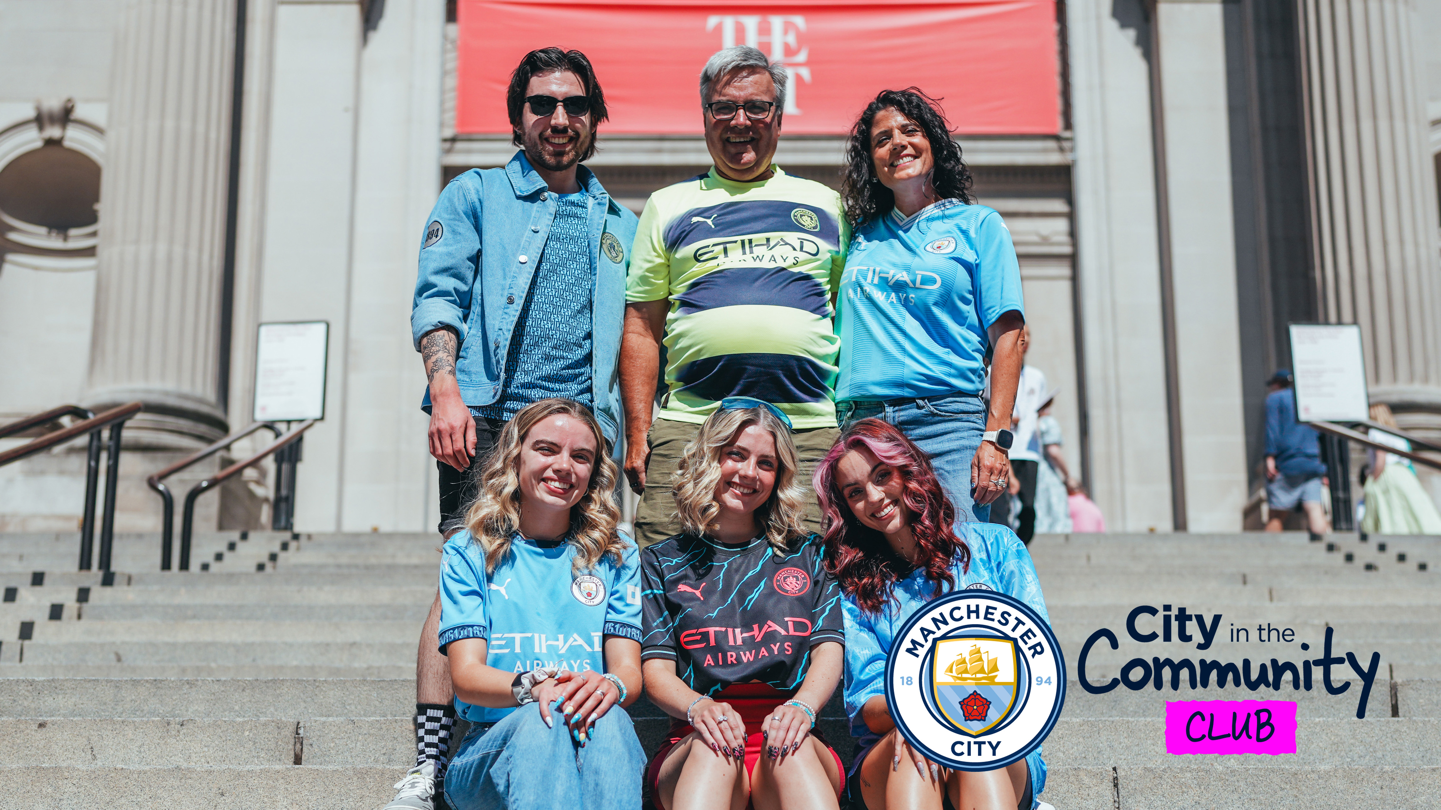 A group of six people wearing Manchester City jerseys, some sitting, some standing on steps with a 'City in the Community' logo.