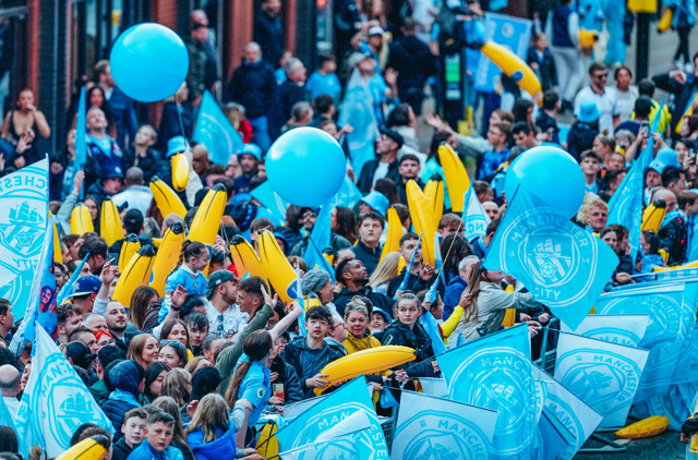 A crowd of Manchester City fans celebrating, holding blue flags and balloons with club logo.