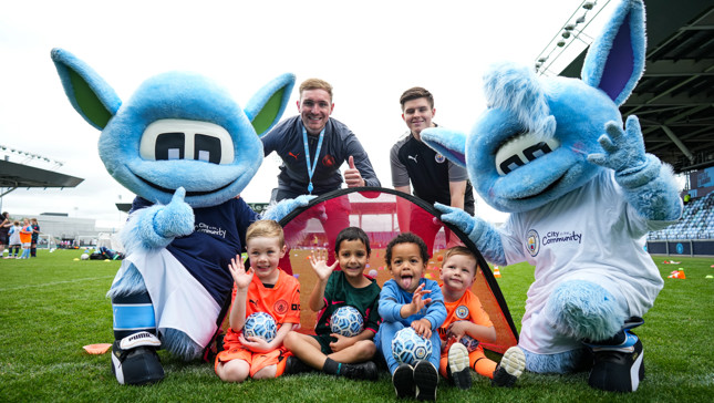 Two mascots pose with children sitting with footballs on a field, alongside two adults behind them.