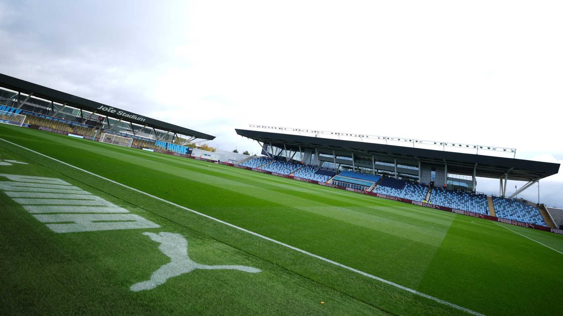 A football stadium with green grass and blue seats, labeled 'Joie Stadium'. The Puma logo is visible on the field.