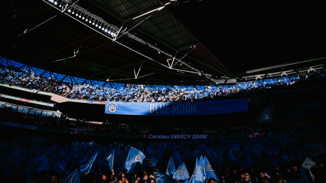 Image of a crowd at a stadium with Manchester City flags and 'Blue Moon' banner.