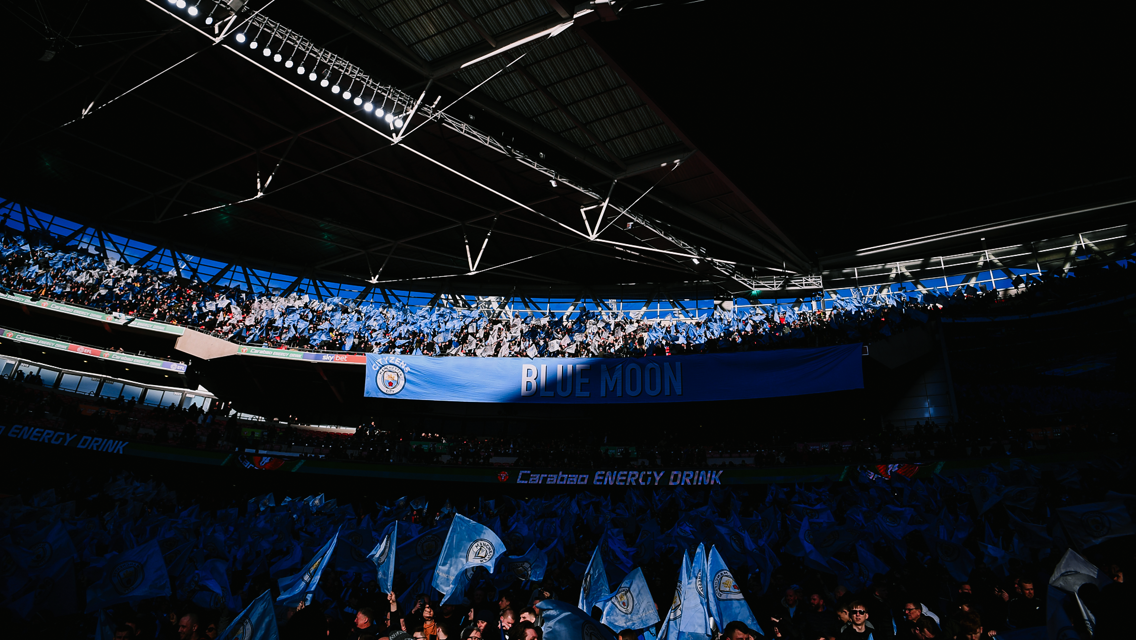Image of a crowd at a stadium with Manchester City flags and 'Blue Moon' banner.