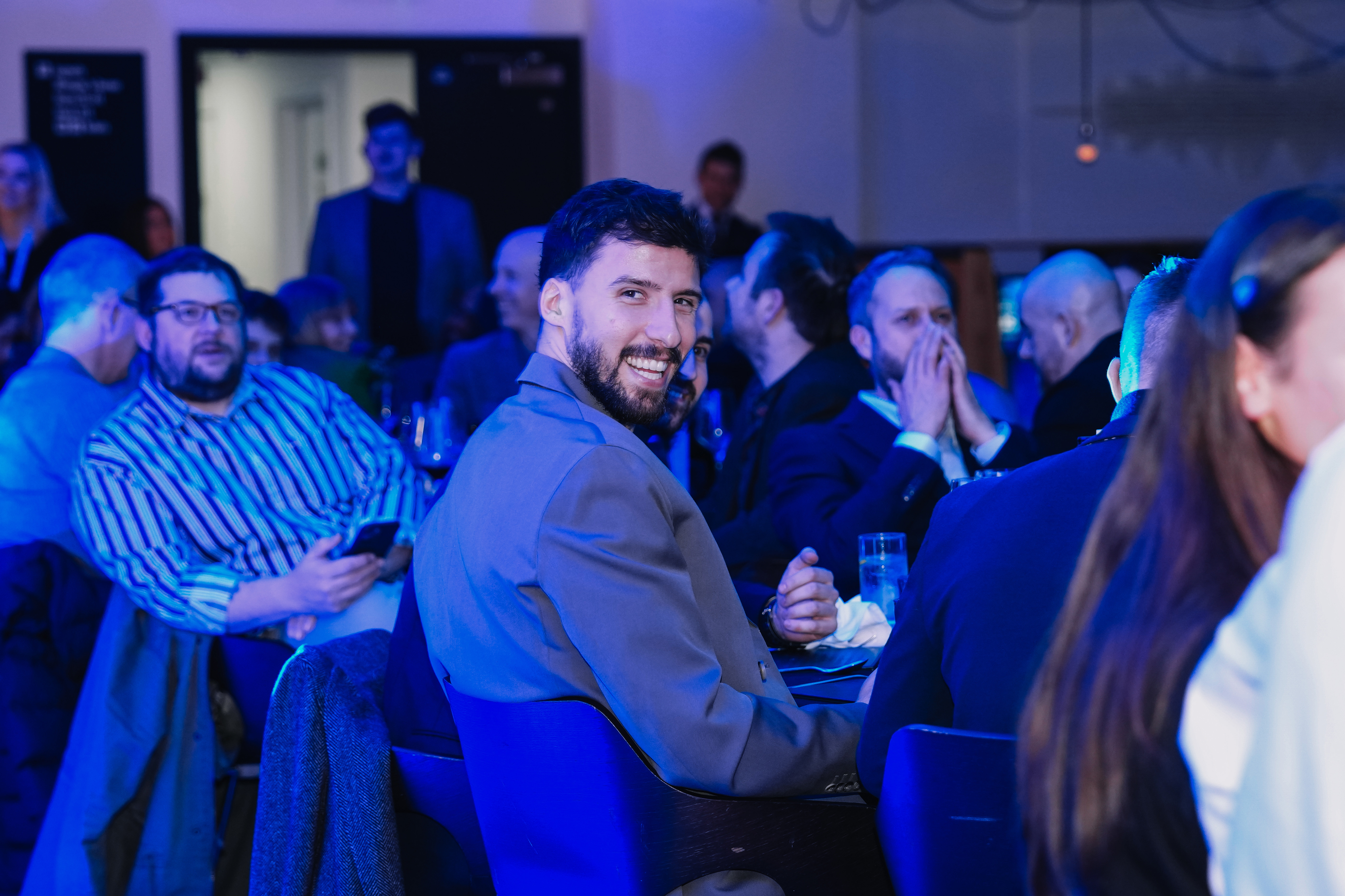 A man in a gray blazer smiling while seated at a table in a crowded room with people in formal attire.