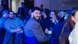 A man in a gray blazer smiling while seated at a table in a crowded room with people in formal attire.