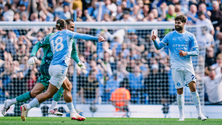 Football players in Manchester City kits celebrate a goal on the field with a crowd of fans in the background.
