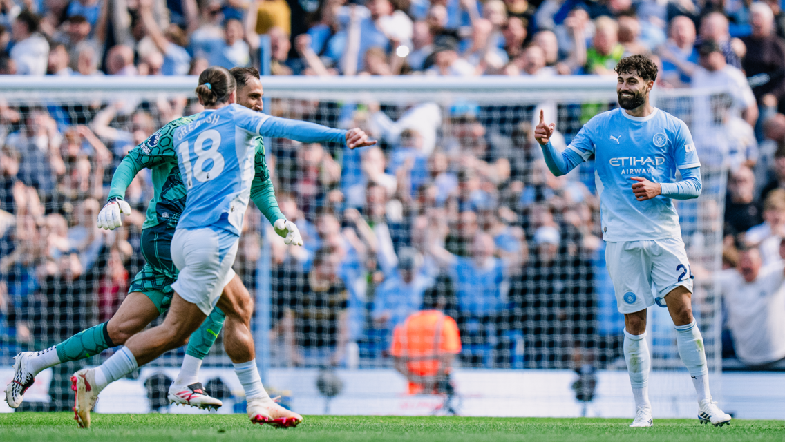 Football players in Manchester City kits celebrate a goal on the field with a crowd of fans in the background.