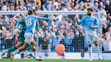 Football players in Manchester City kits celebrate a goal on the field with a crowd of fans in the background.