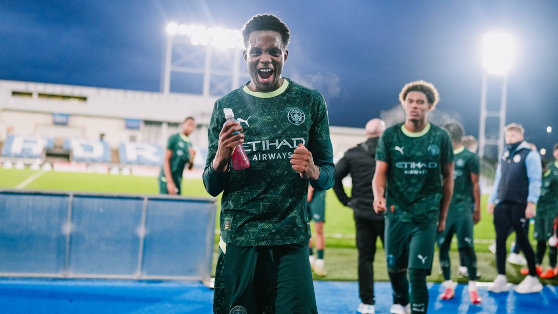Several young athletes in Manchester City green training kits walking with hydration bottles in hand on a football field under bright floodlights.