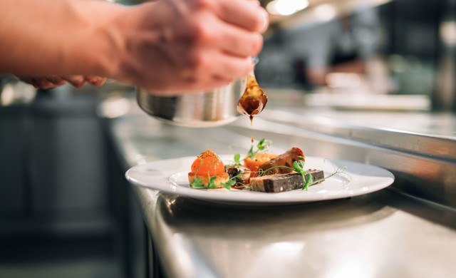 A chef carefully garnishing a plated dish with sauce using a spoon in a professional kitchen setting.
